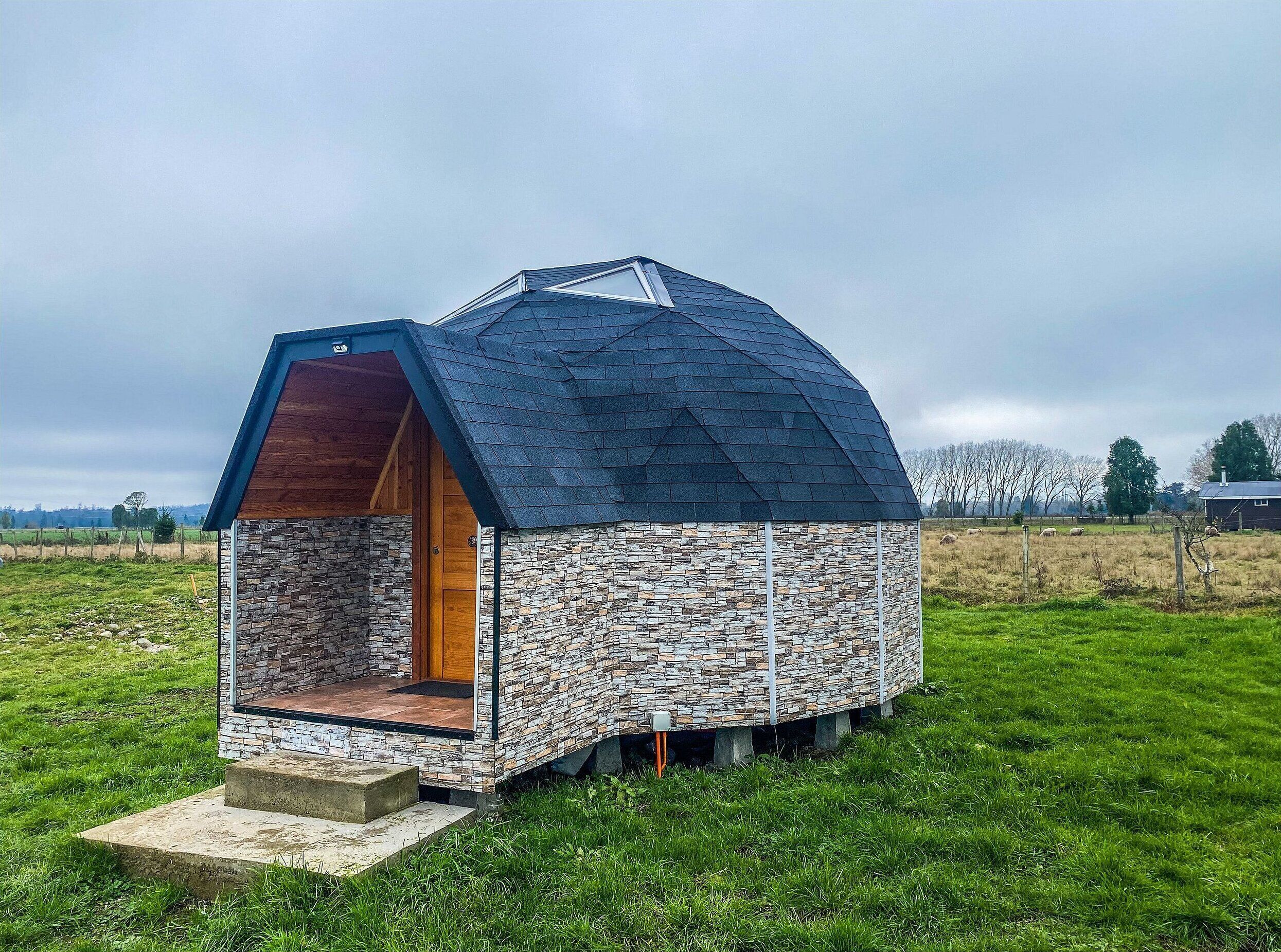A small, stone-clad structure with a sloped roof and a wooden door, situated on a grassy field.