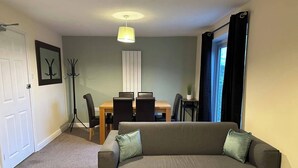 Ein Wohnzimmer mit grauem Sofa, einem Holztisch, einem Esstisch mit Stühlen, einer Stehlampe und einem Fenster mit Vorhängen.