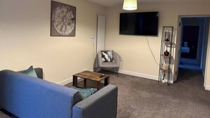 Ein Wohnzimmer mit einem blauen Sofa, einem kleinen Holztisch, einem grauen Sessel, einem an der Wand befestigten Fernseher und einer Deckenleuchte.