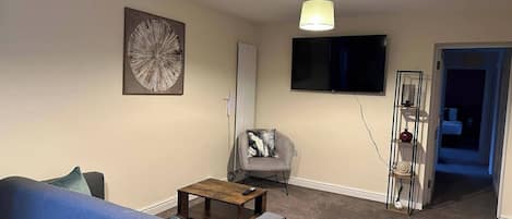A living room with a blue sofa, a small wooden table, a gray armchair, a TV mounted on the wall, and a ceiling light fixture.