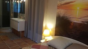A bedroom with a bed, a nightstand with a lamp, a shower area, and a wall with a sunset painting.