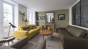 A modern living room with a yellow sofa, a brown sofa, and a round coffee table. There is a fireplace with a mirror above it, and a large window on the right side. - A CENTRAL PARISIAN EXPERIENCE WITH MODERN CONVENIENCES IN THE ICONIC LE MARAIS (Paris)