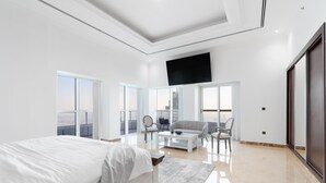 A modern hotel room with a large bed, a flat-screen TV mounted on the wall, a balcony with a city view, and a seating area with chairs and a table. - 4BR Maid Penthouse w Pool and Jacuzzi (Dubai)