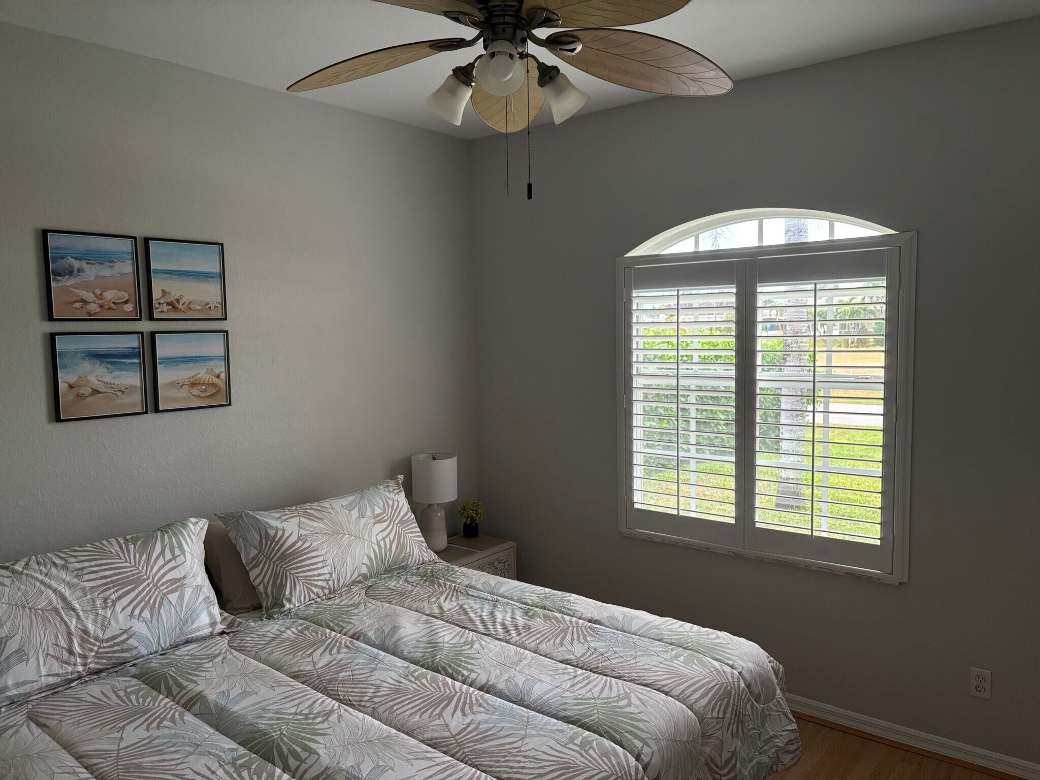 Un dormitorio con cama, ventilador de techo, ventana con persianas y cuatro cuadros en la pared.