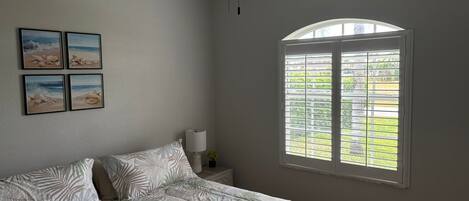 Un dormitorio con cama, ventilador de techo, ventana con persianas y cuatro cuadros en la pared.