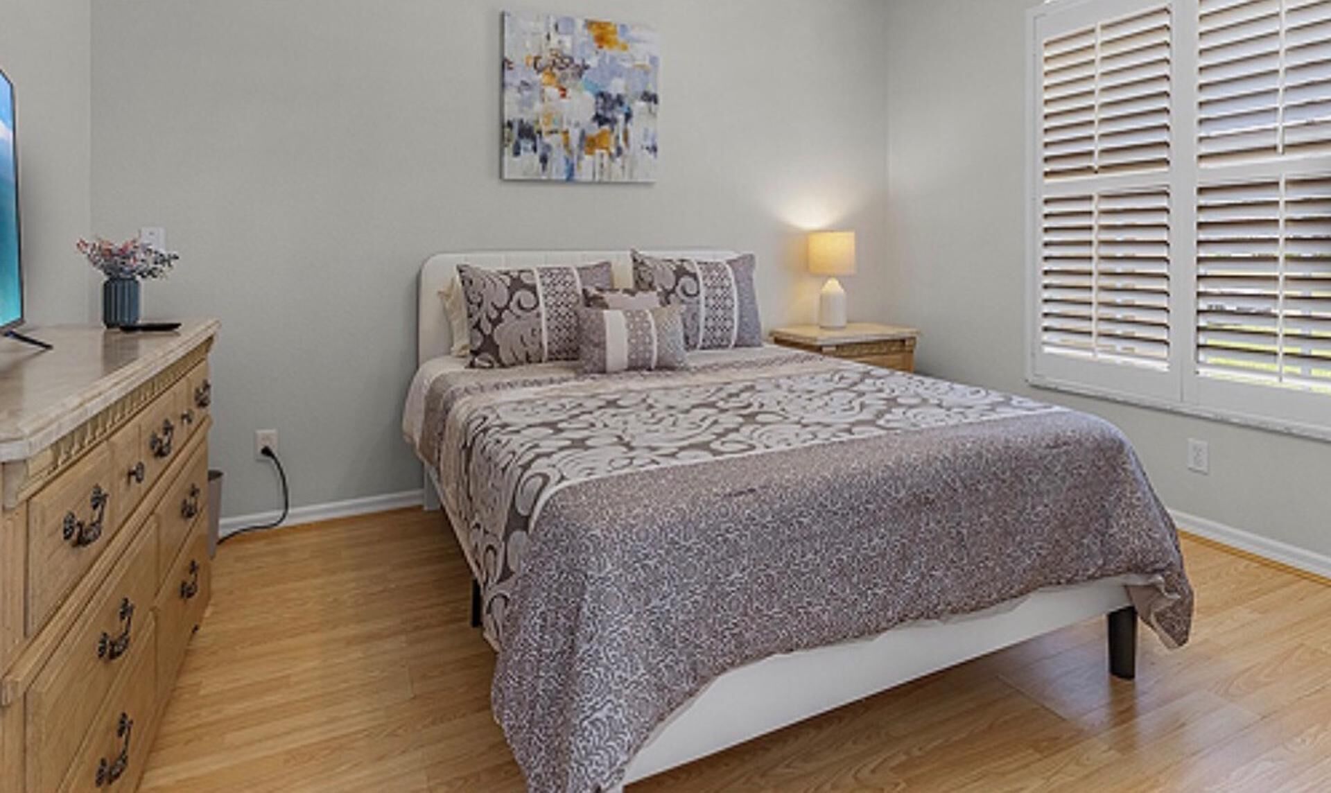 A bedroom with a bed, dresser, nightstand, lamp, and a window with blinds.