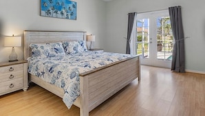 Quarto com cama de madeira, mesa de cabeceira, luminária e pintura de palmeiras.