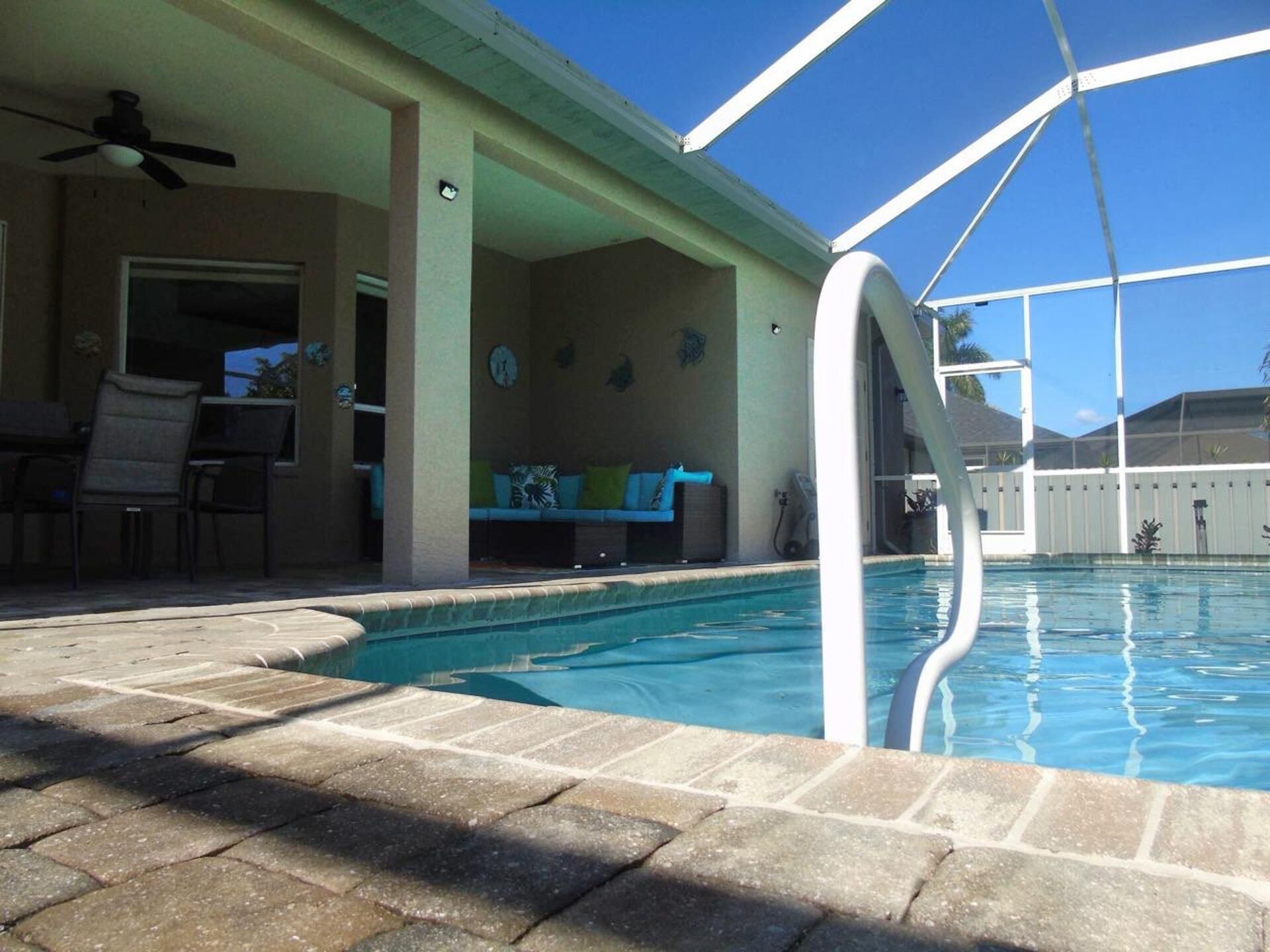 A swimming pool with a covered patio area, a ceiling fan, and a seating area with a couch and chairs.