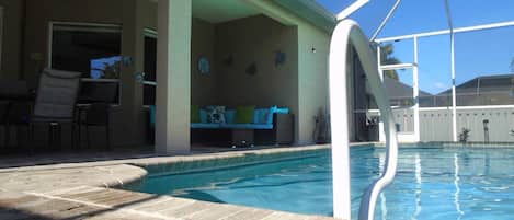 A swimming pool with a covered patio area, a ceiling fan, and a seating area with a couch and chairs.