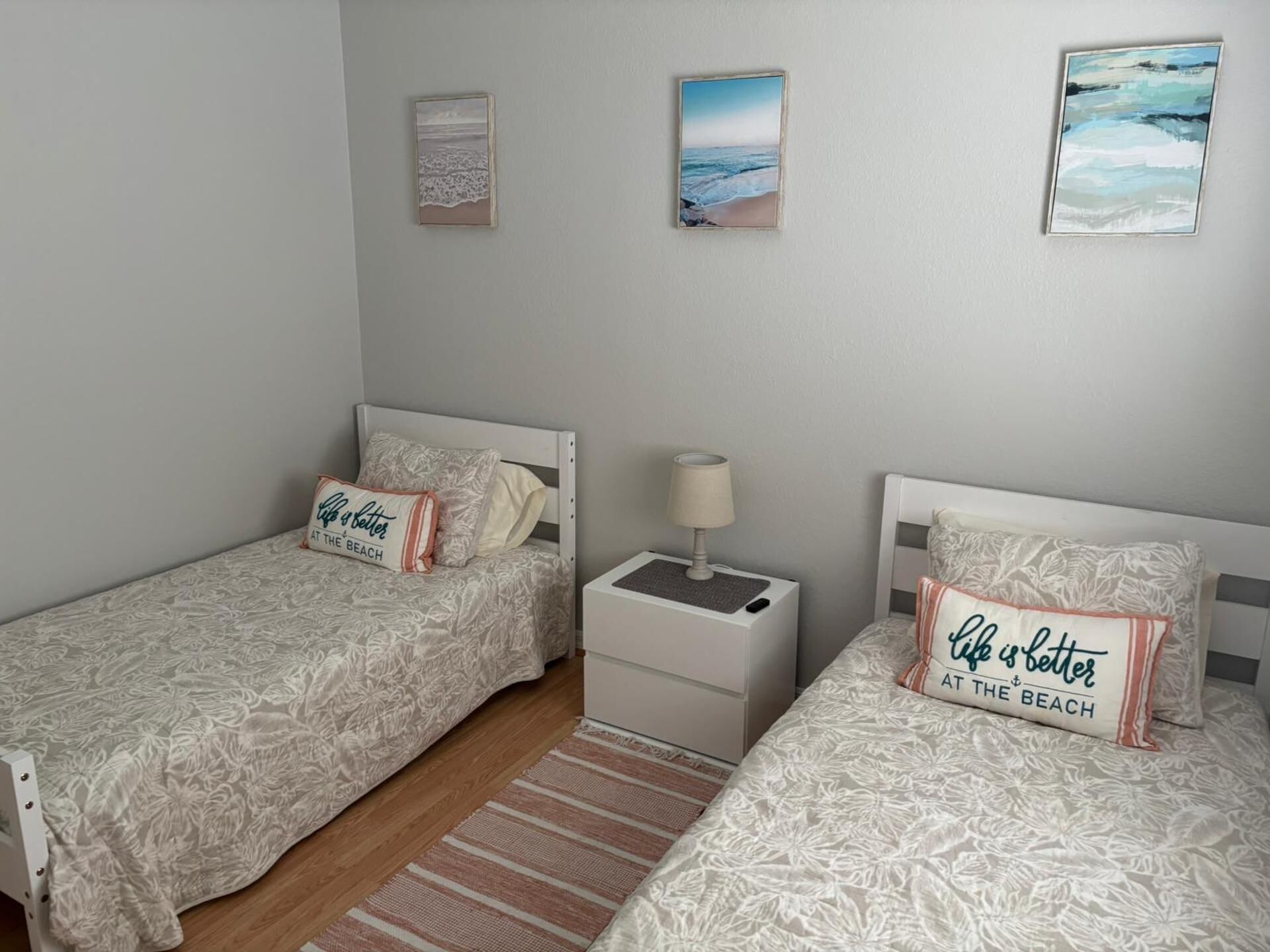 Ein Schlafzimmer mit zwei Betten, jedes mit einem Kissen, auf dem steht 'Life is better at the beach'. Zwei Bilder hängen an der Wand, eines zeigt einen Strand und das andere eine Stadtansicht.