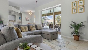 Uma sala de estar moderna com um sofá seccional cinza, uma mesa de centro redonda com uma planta e vista para um pátio com móveis de exterior.