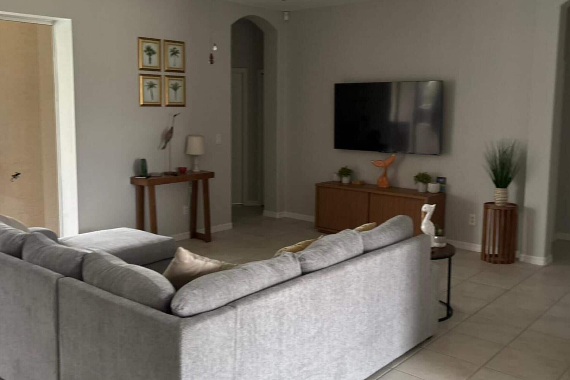 A living room with a grey sofa, a television mounted on the wall, and a small table with a lamp.