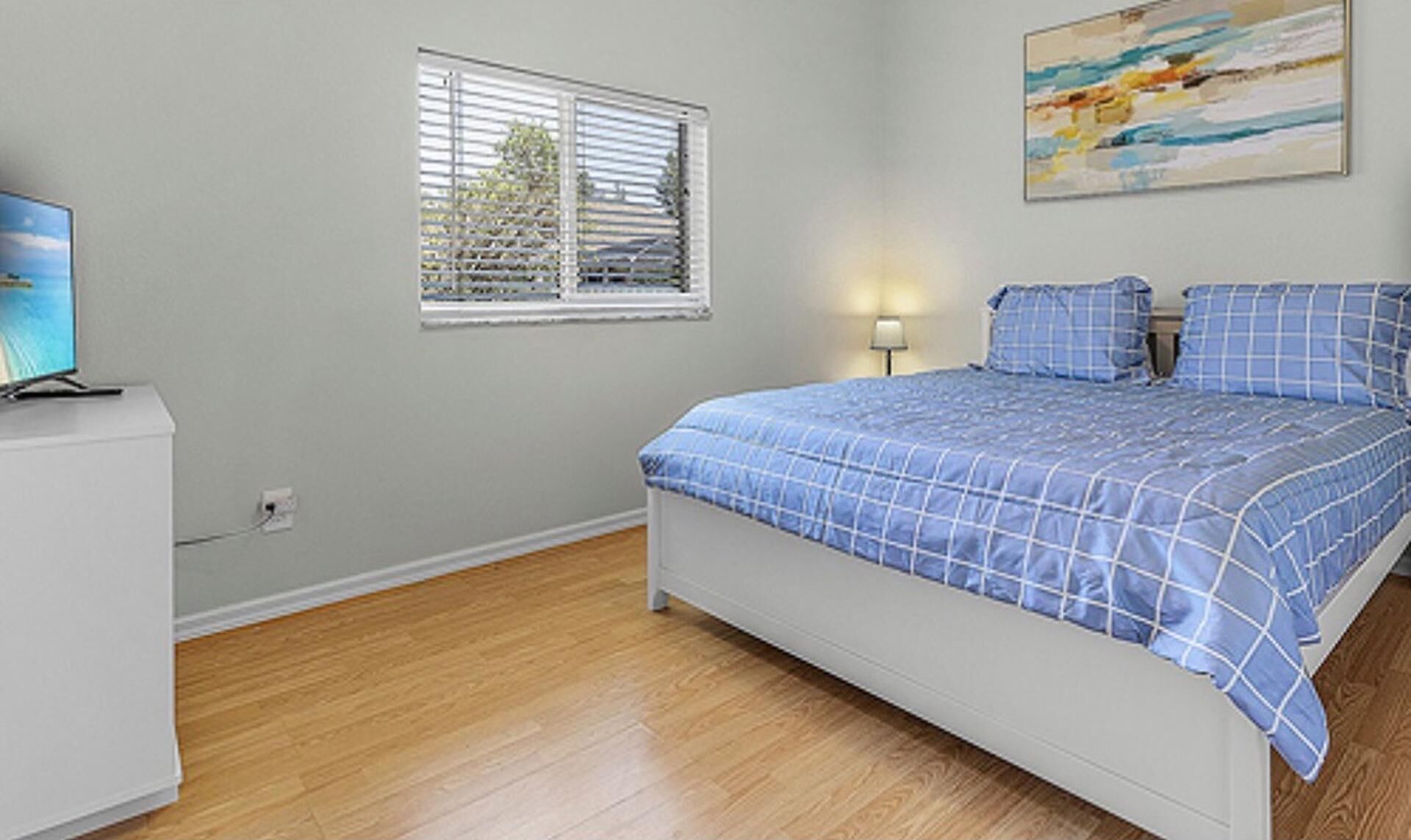 Un dormitorio con cama, ventana con persianas, un cuadro en la pared y un televisor sobre un mueble.