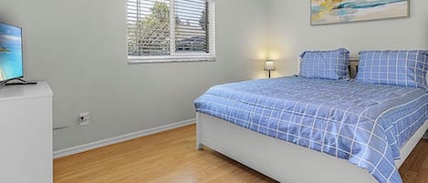 Ein Schlafzimmer mit Bett, einem Fenster mit Jalousien, einem Bild an der Wand und einem Fernseher auf einem Schrank.