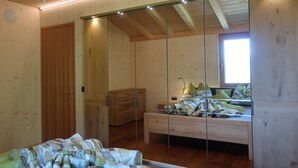A bedroom with a wooden floor, a bed with a patterned bedspread, a bedside table, a mirror, and a window. - Rumwaldhof Edelweiss 82-m² apartment with unique view of Ortles (Sulden)