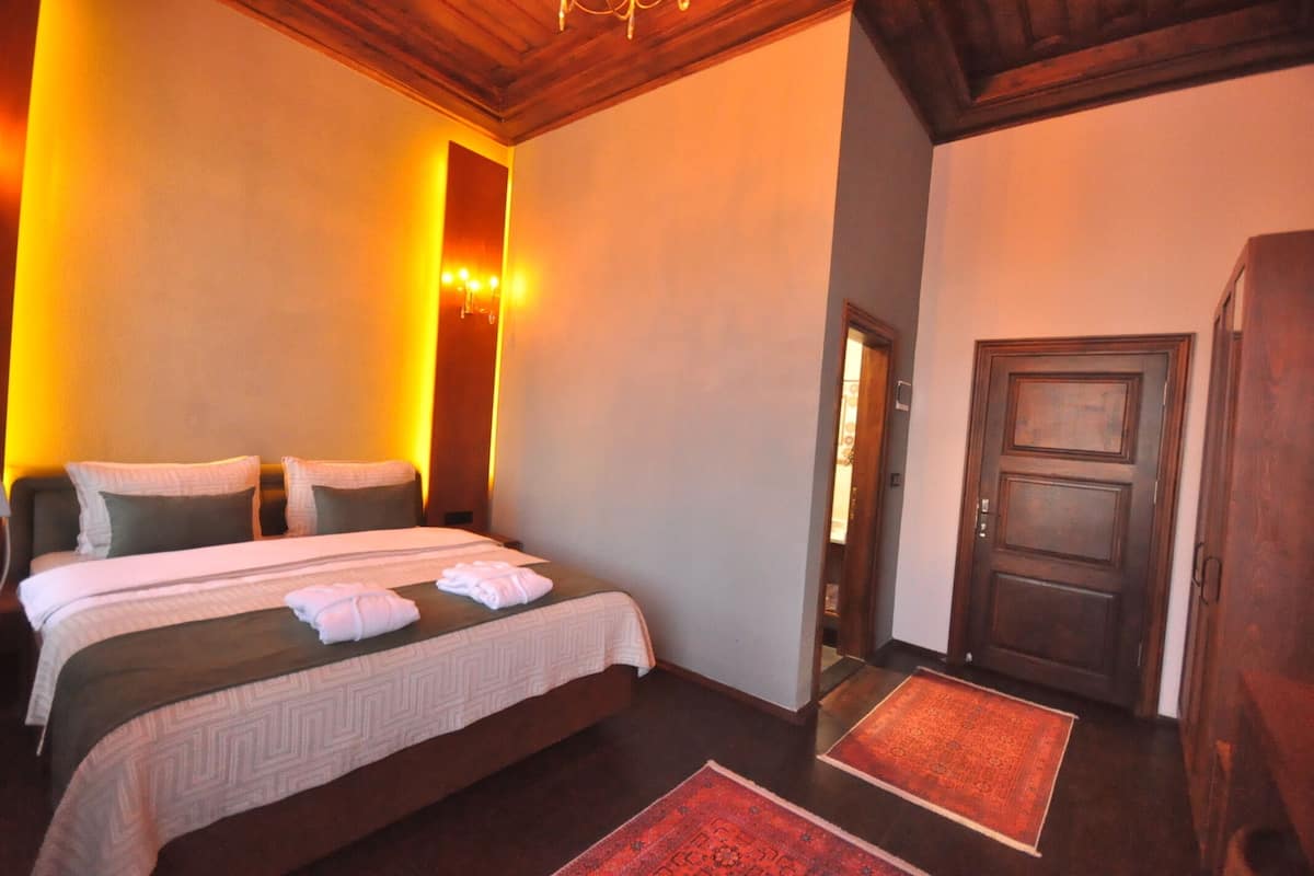 Quarto de hotel com uma cama grande, duas almofadas, uma mesa de cabeceira, um tapete vermelho e uma porta de madeira.