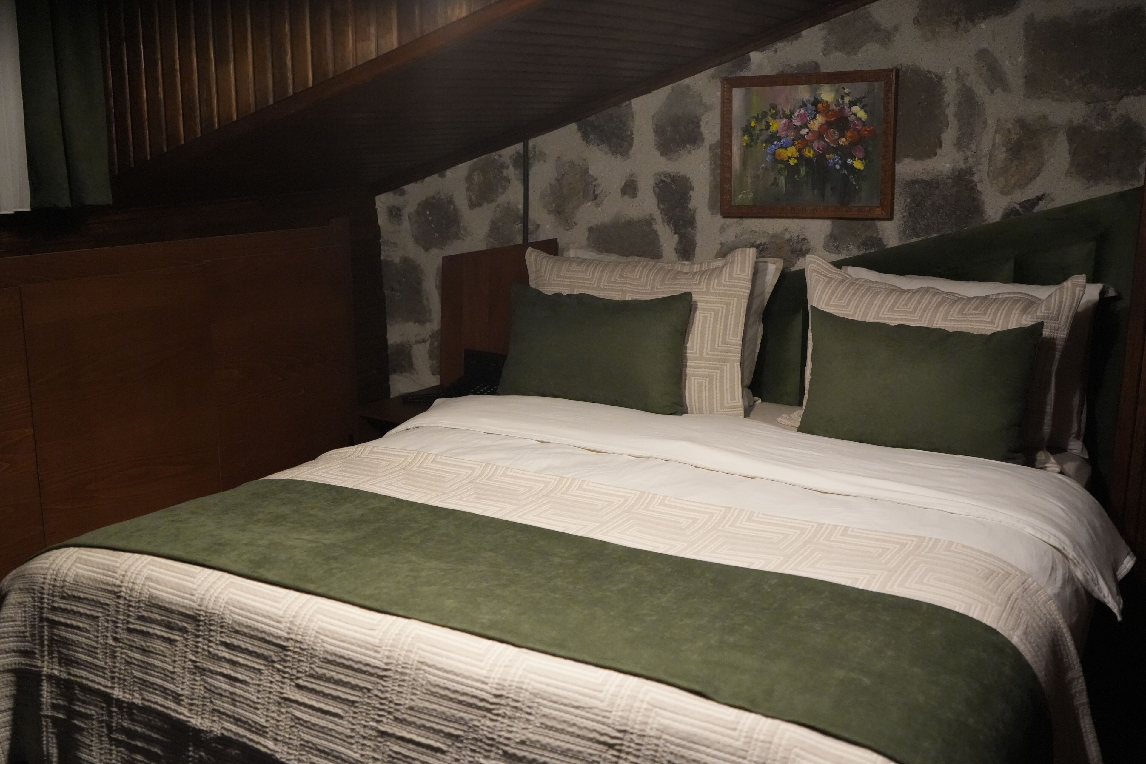 A bedroom with a stone wall, a wooden headboard, a green bedspread, and a painting of flowers on the wall.
