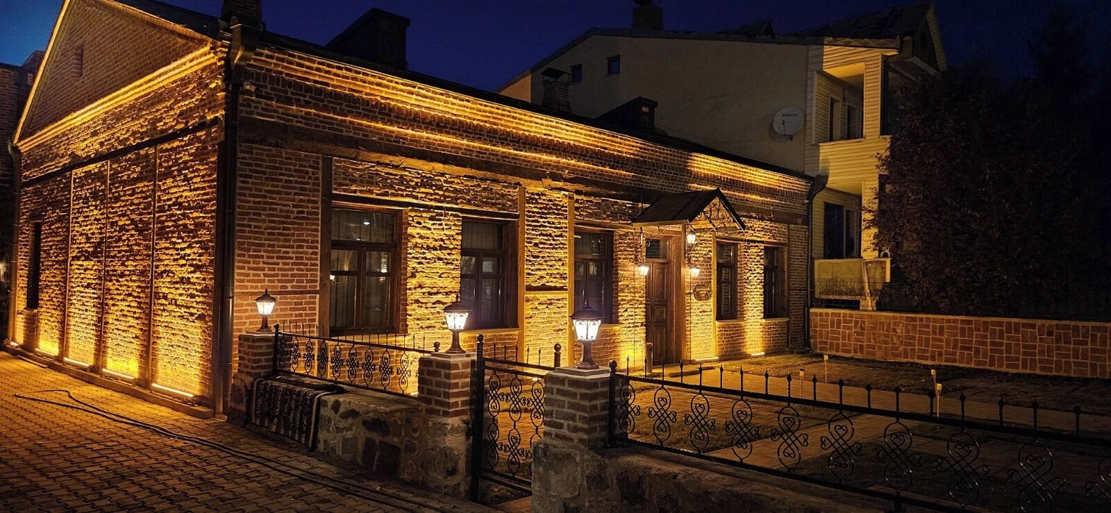 Un edificio de piedra con fachada de ladrillo, iluminado con luz cálida, que presenta una barandilla metálica y un acceso empedrado.