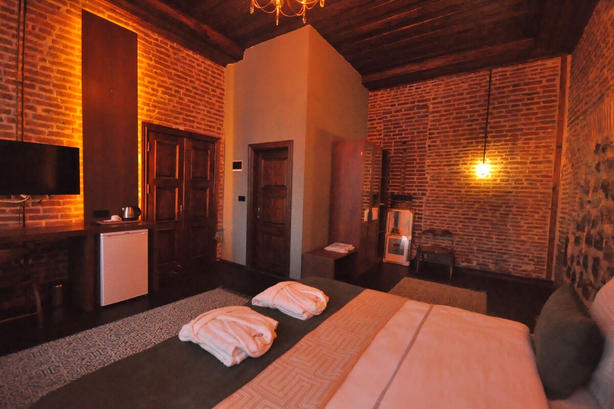 A hotel room with a brick wall, a bed with two pillows, a small table with a kettle, a TV, and a refrigerator.