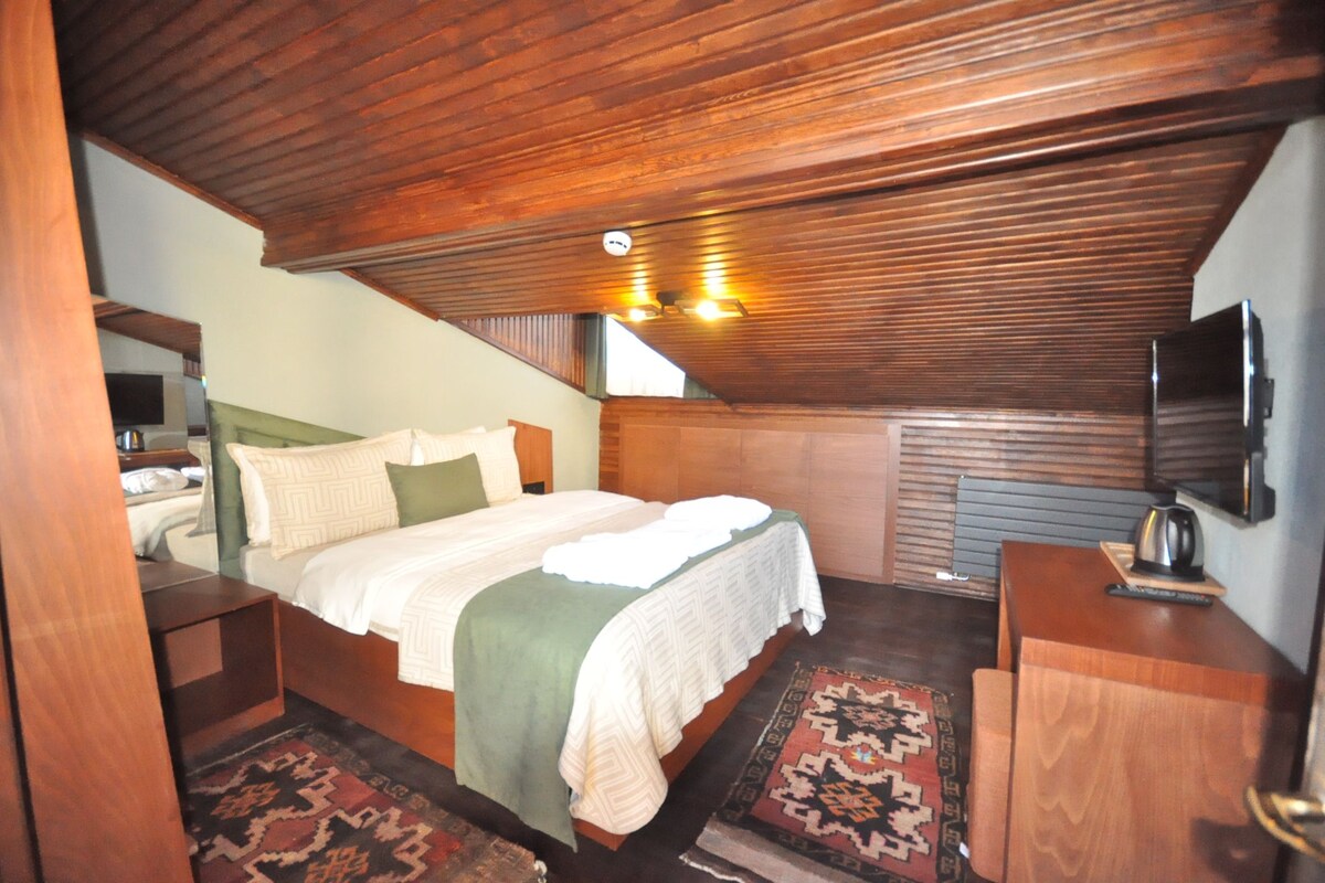 A bedroom with a wooden ceiling, a bed with a green and white bedspread, a nightstand, a dresser, a television mounted on the wall, and a patterned rug on the floor.