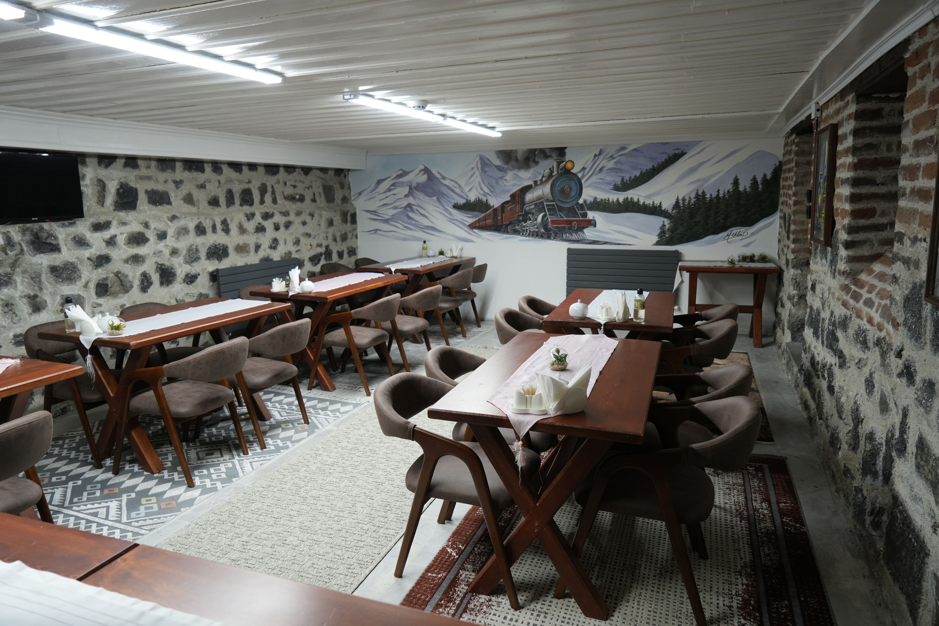 Un espace repas avec des tables et des chaises en bois, un mur en pierre et une fresque représentant un train dans une montagne.