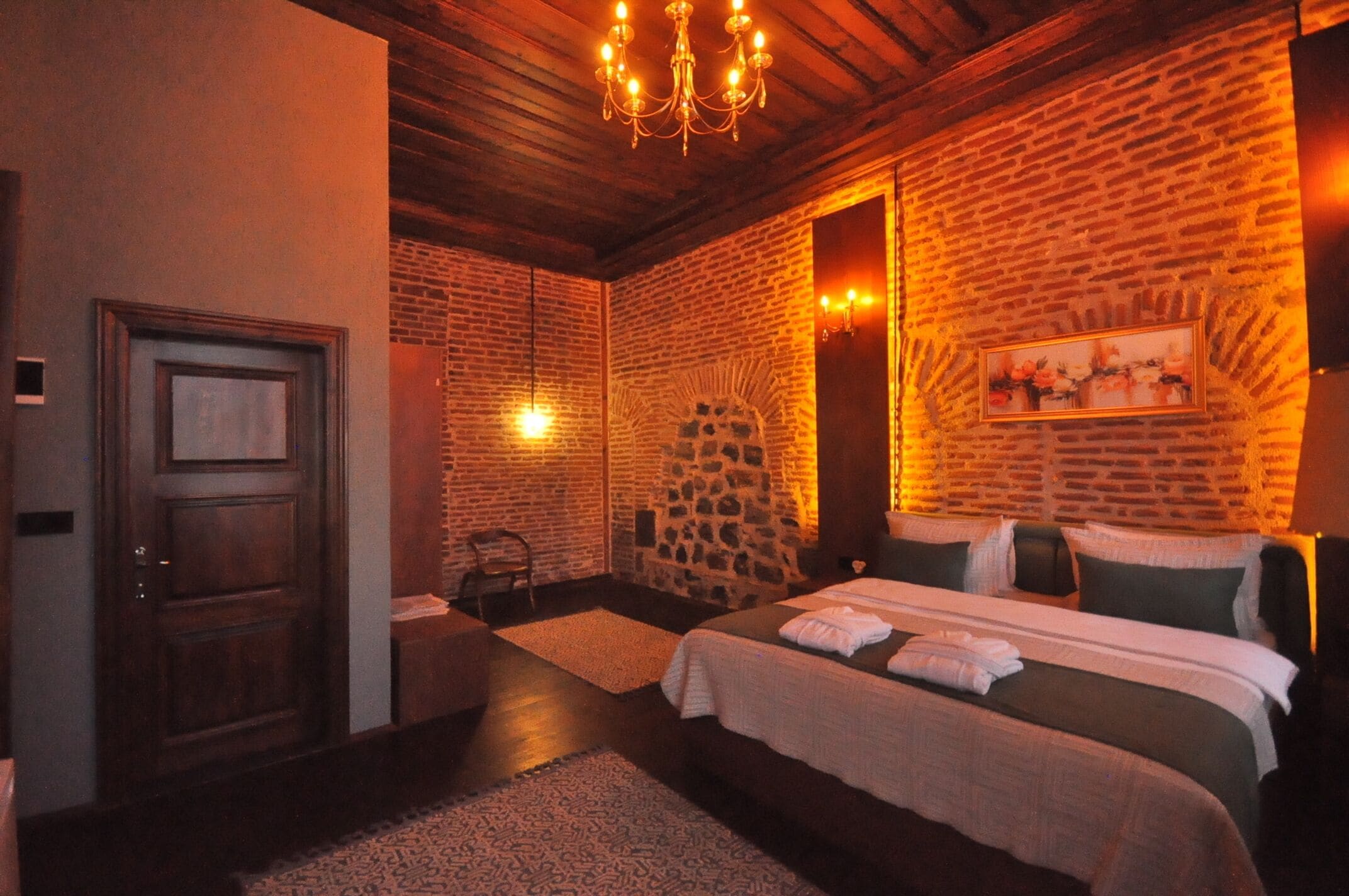 A hotel room with a brick wall, a chandelier, a bed with pillows, and a door.