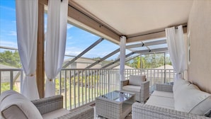 A covered outdoor seating area with wicker furniture and a glass table, overlooking a garden. - Unique, 7 bed Luxury Disney Escape, Theater & Heated Pool (Davenport)