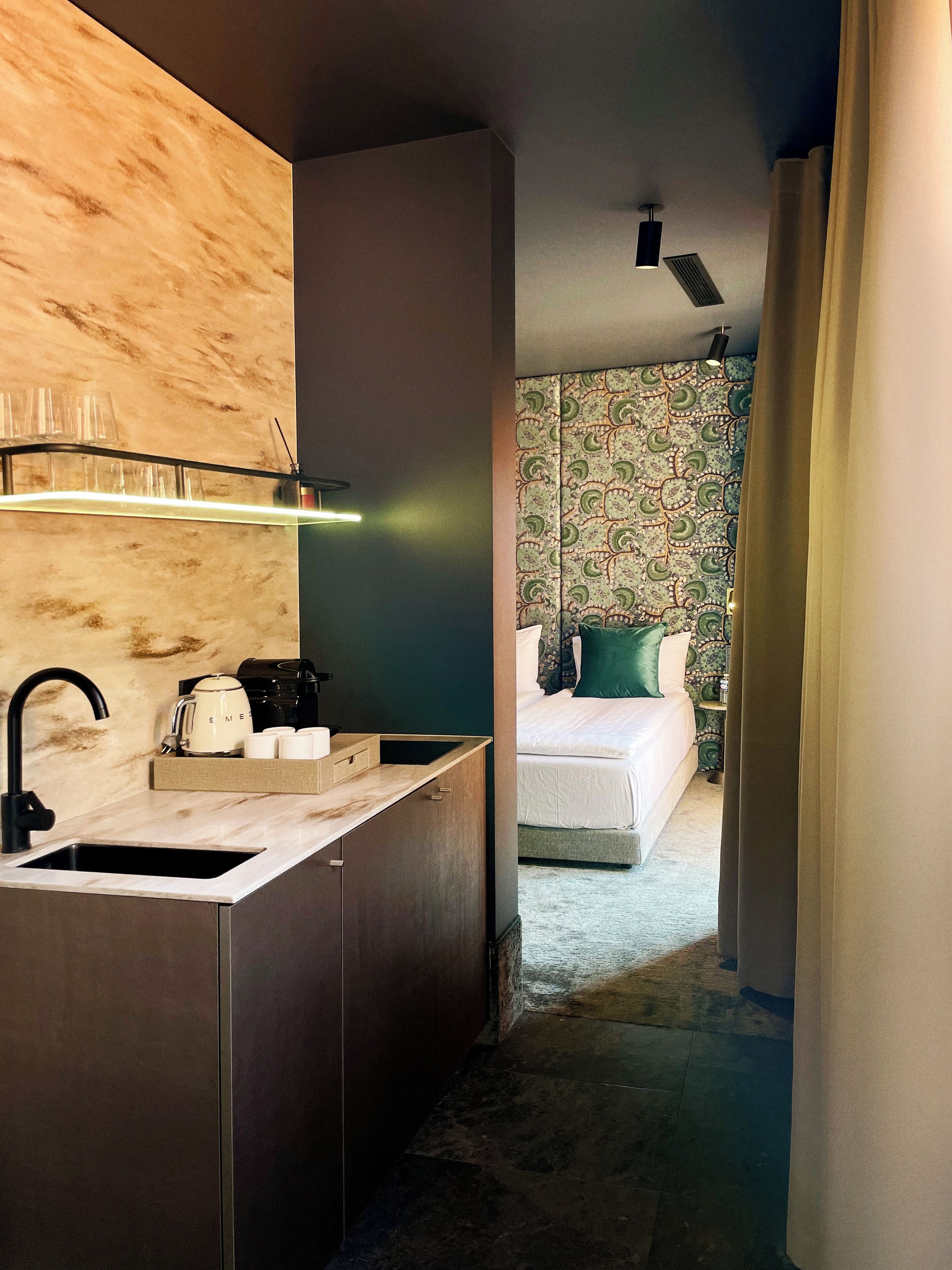 A modern hotel room with a kitchen area, a bed with a patterned headboard, and a view of the hallway.