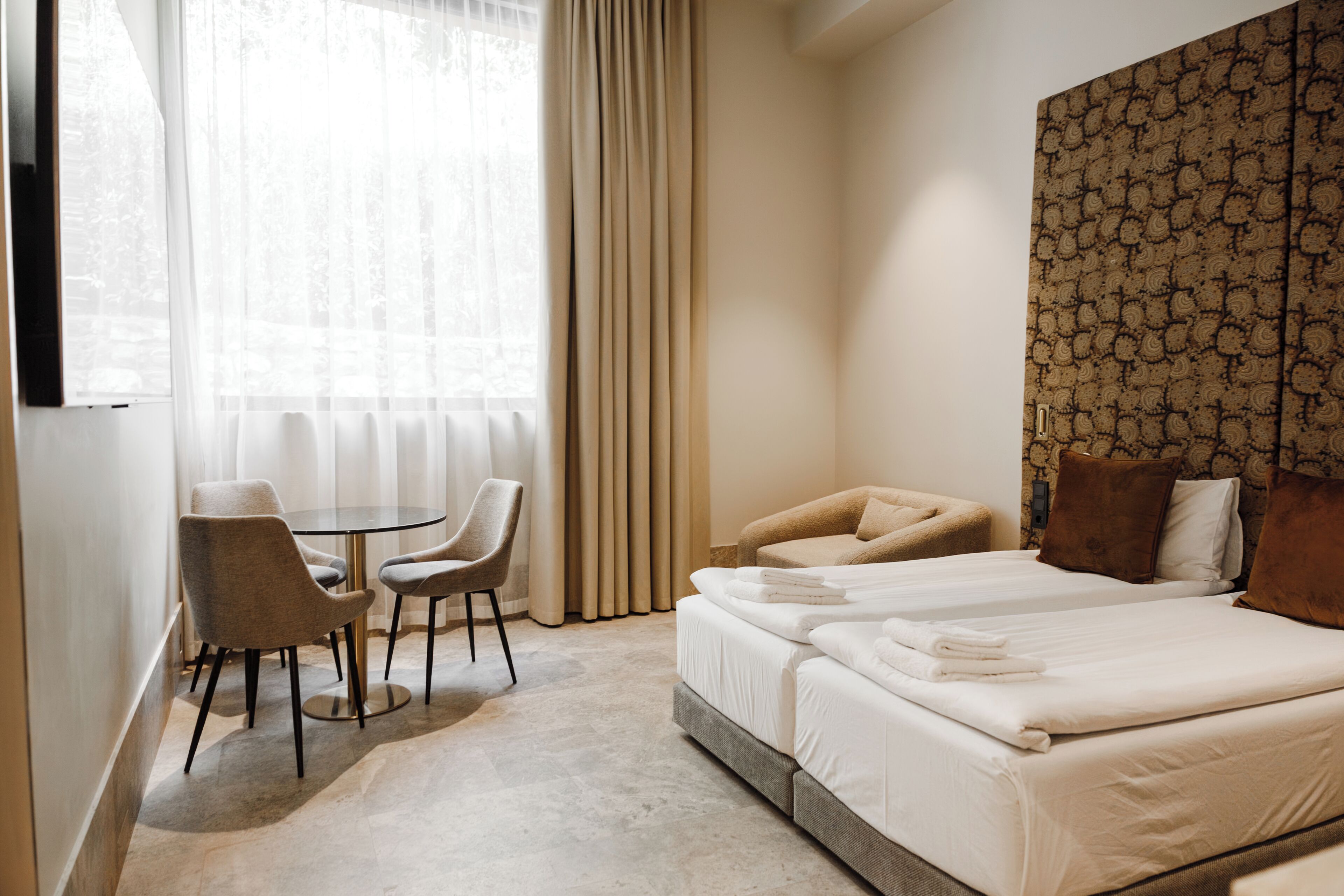 Une chambre d’hôtel avec un lit, deux chaises, une petite table et une grande fenêtre avec des rideaux.