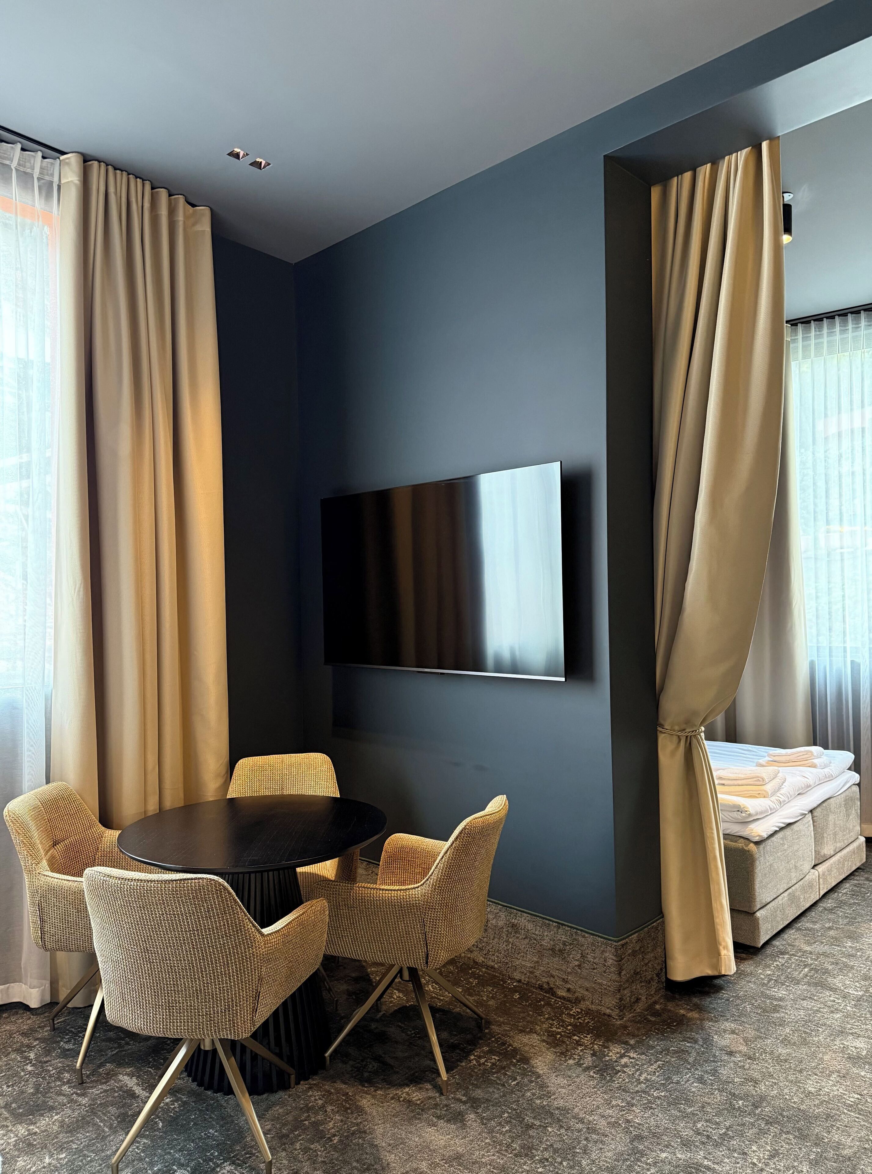 Een moderne kamer met een ronde tafel en stoelen, een flatscreen televisie aan de muur en een bed met gordijnen.