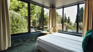 Une chambre avec une grande fenêtre, un lit, une chaise et une petite table sur laquelle se trouve un vase.
