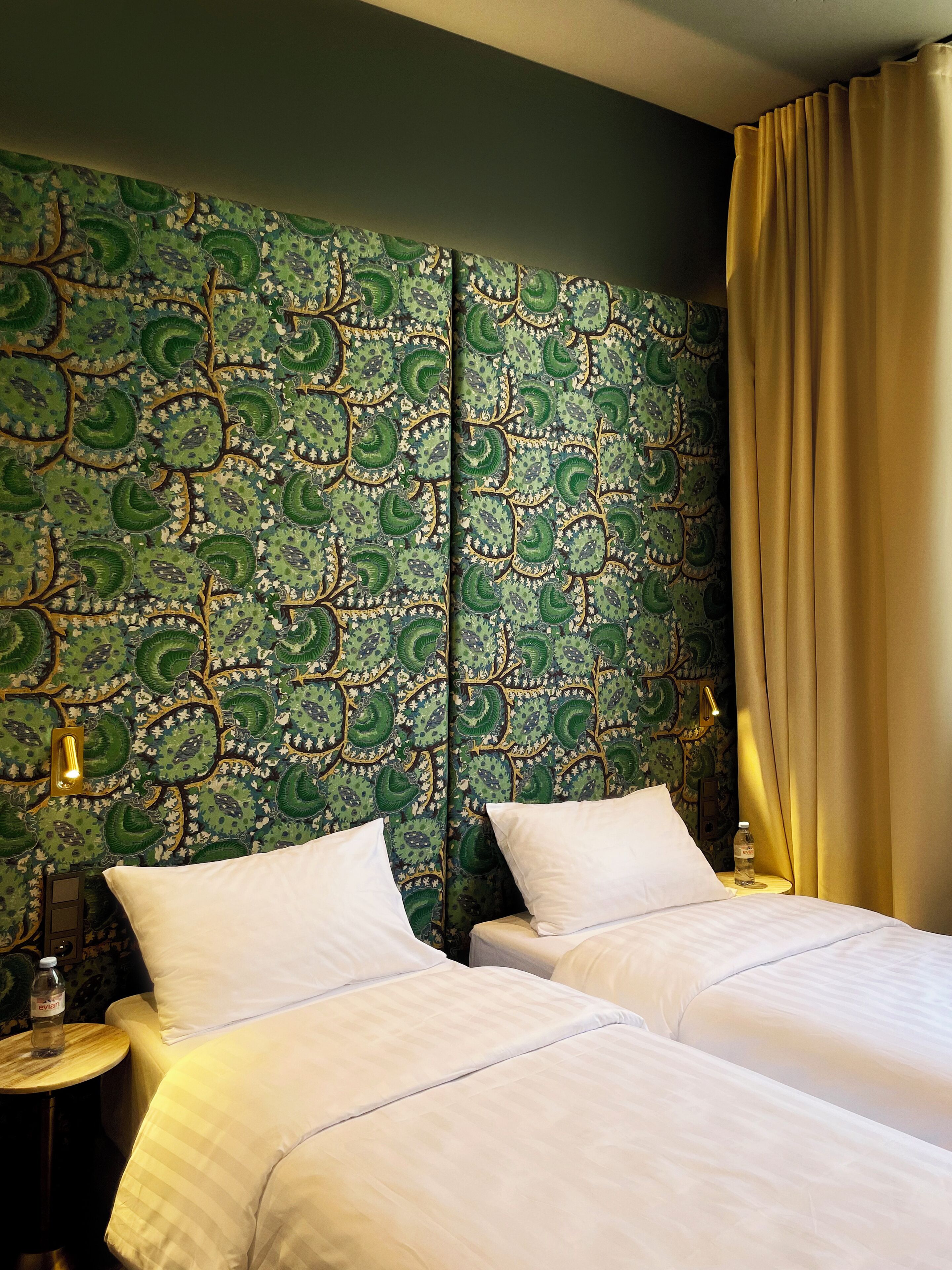 A hotel room with two beds, a green patterned headboard, a round side table with a bottle, and a lamp.