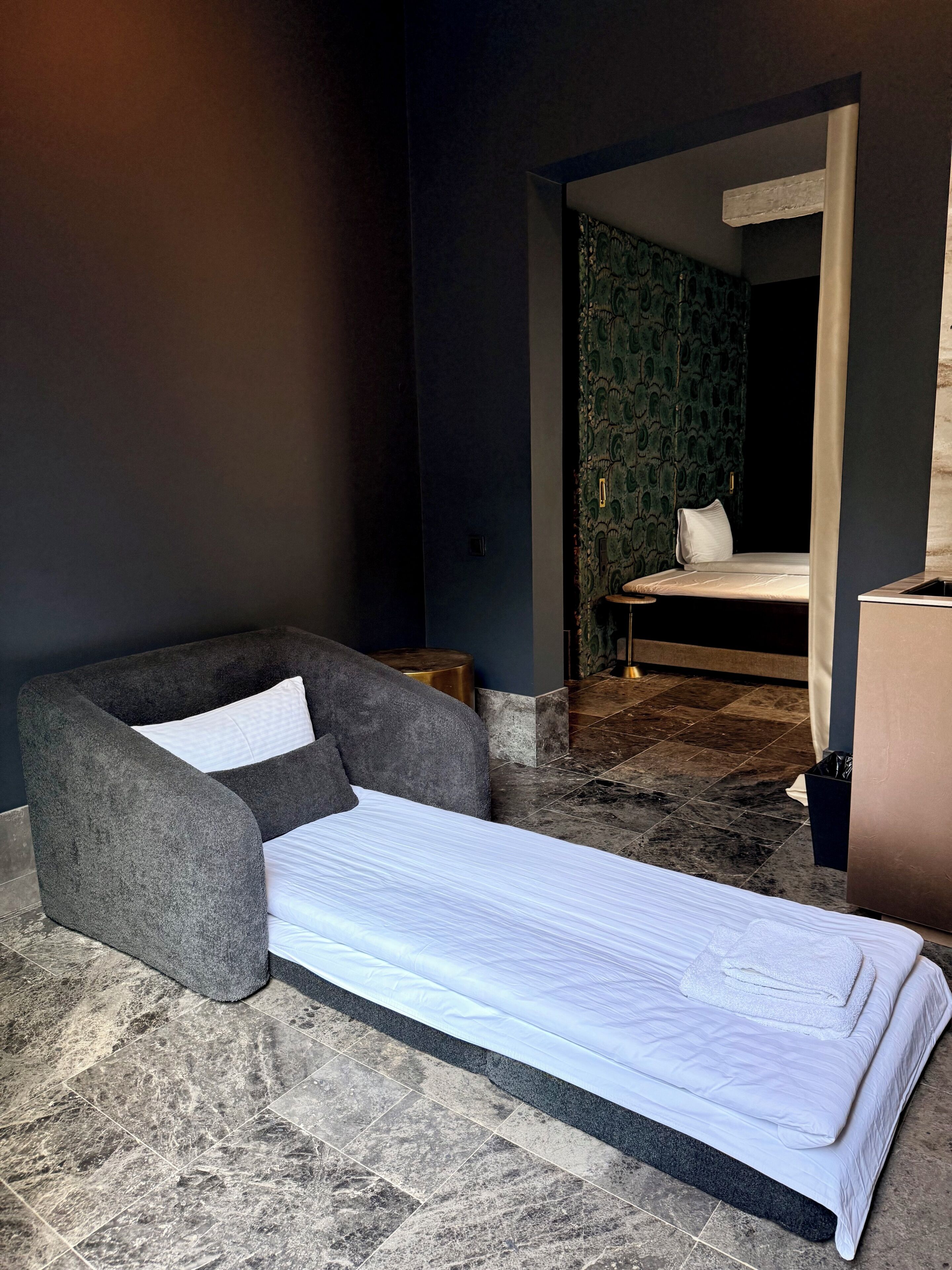 Une chambre avec un lit simple, un fauteuil gris, une petite table et un mur sombre recouvert d’un papier peint à motifs.