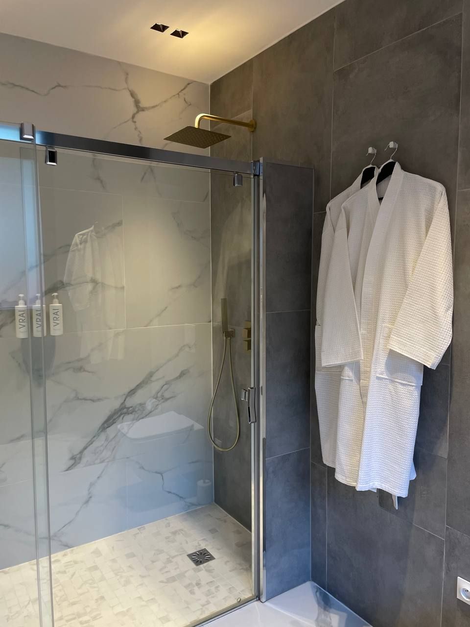 A modern bathroom with a glass-enclosed shower, marble tiles, and a towel rack with robes.