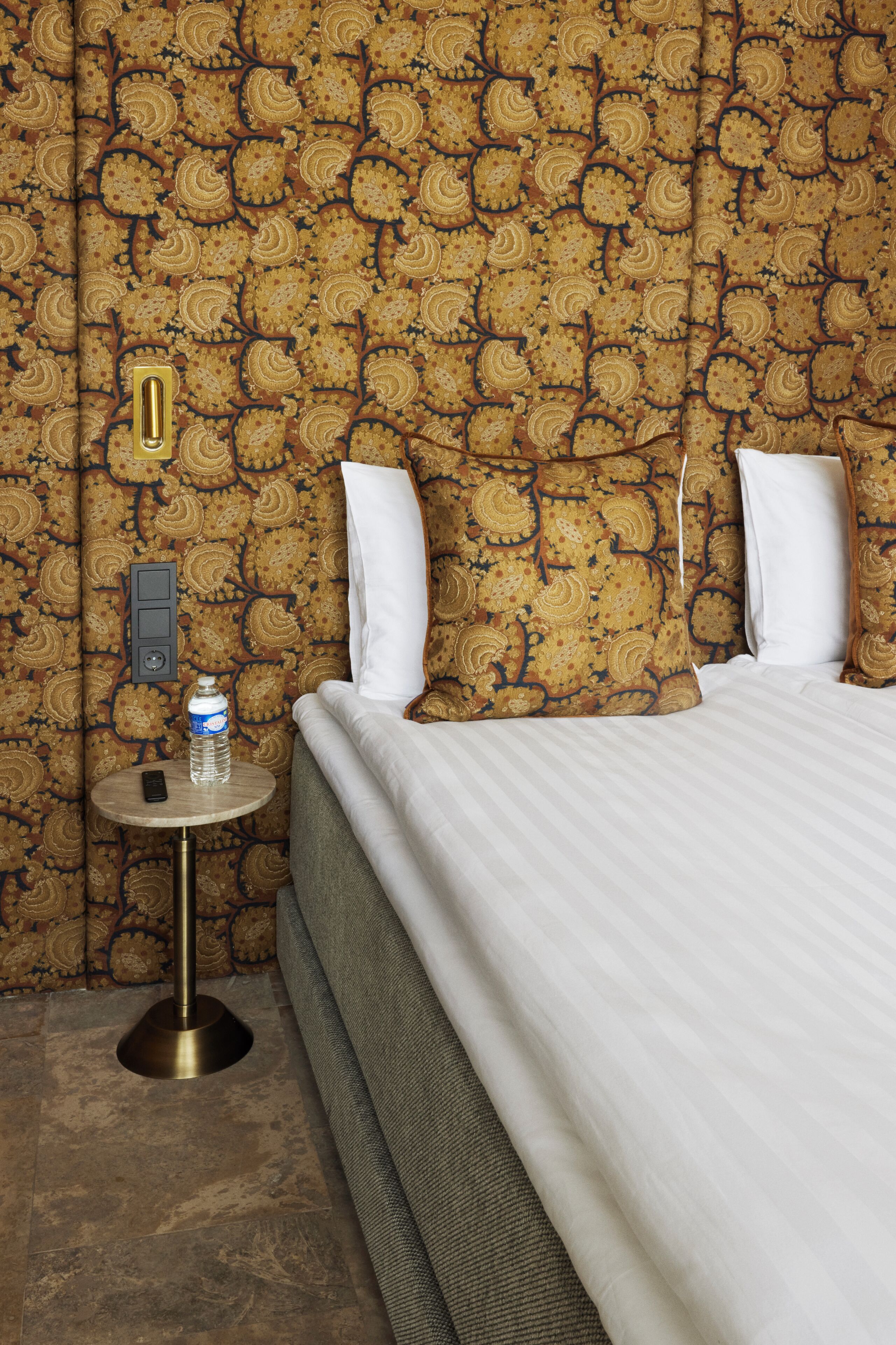 Habitación de hotel con papel tapiz estampado, una cama con ropa blanca, una mesa redonda con una botella y un teléfono, y un interruptor de luz en la pared.