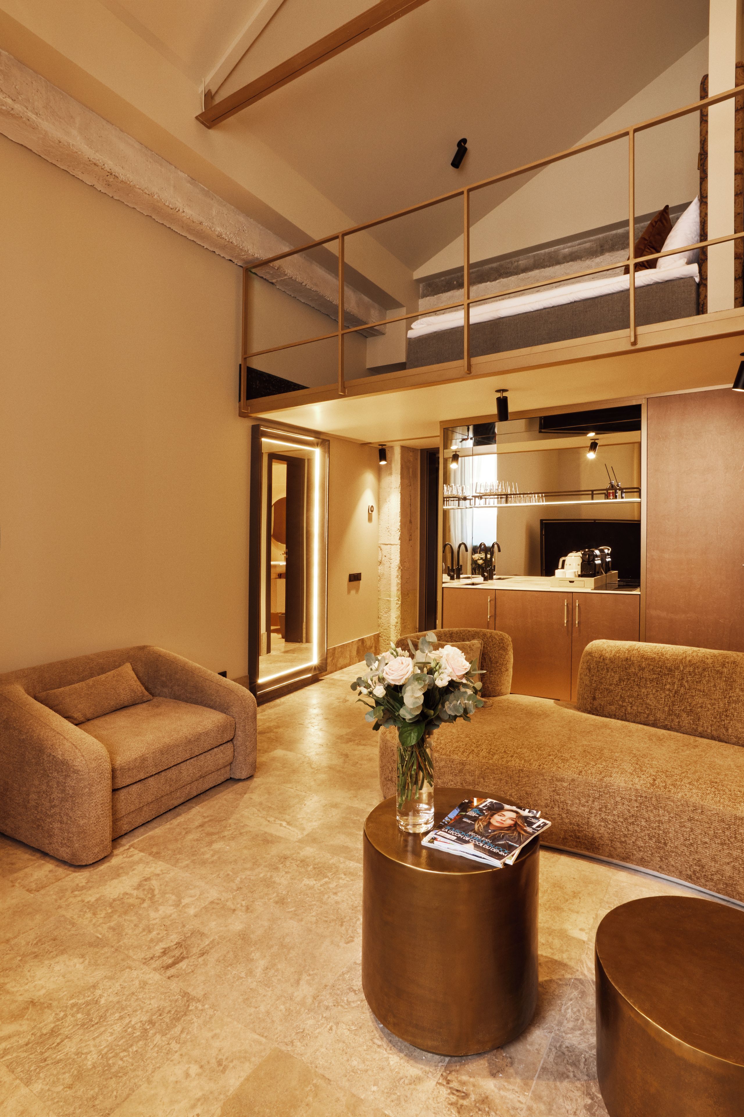 A modern living room with a sofa, a coffee table with a vase and magazine, a kitchen area with a counter and cabinets, and a staircase leading to an upper level.