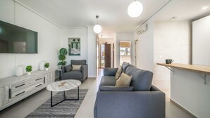 Un soggiorno moderno con un divano grigio, un tavolino da caffè e una TV fissata alla parete. Si nota una mensola in legno con una tazza sopra, e una pianta in vaso sul pavimento.