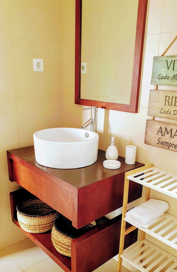 A bathroom with a round white sink, a wooden vanity, a mirror, and a shelf with baskets and a towel.