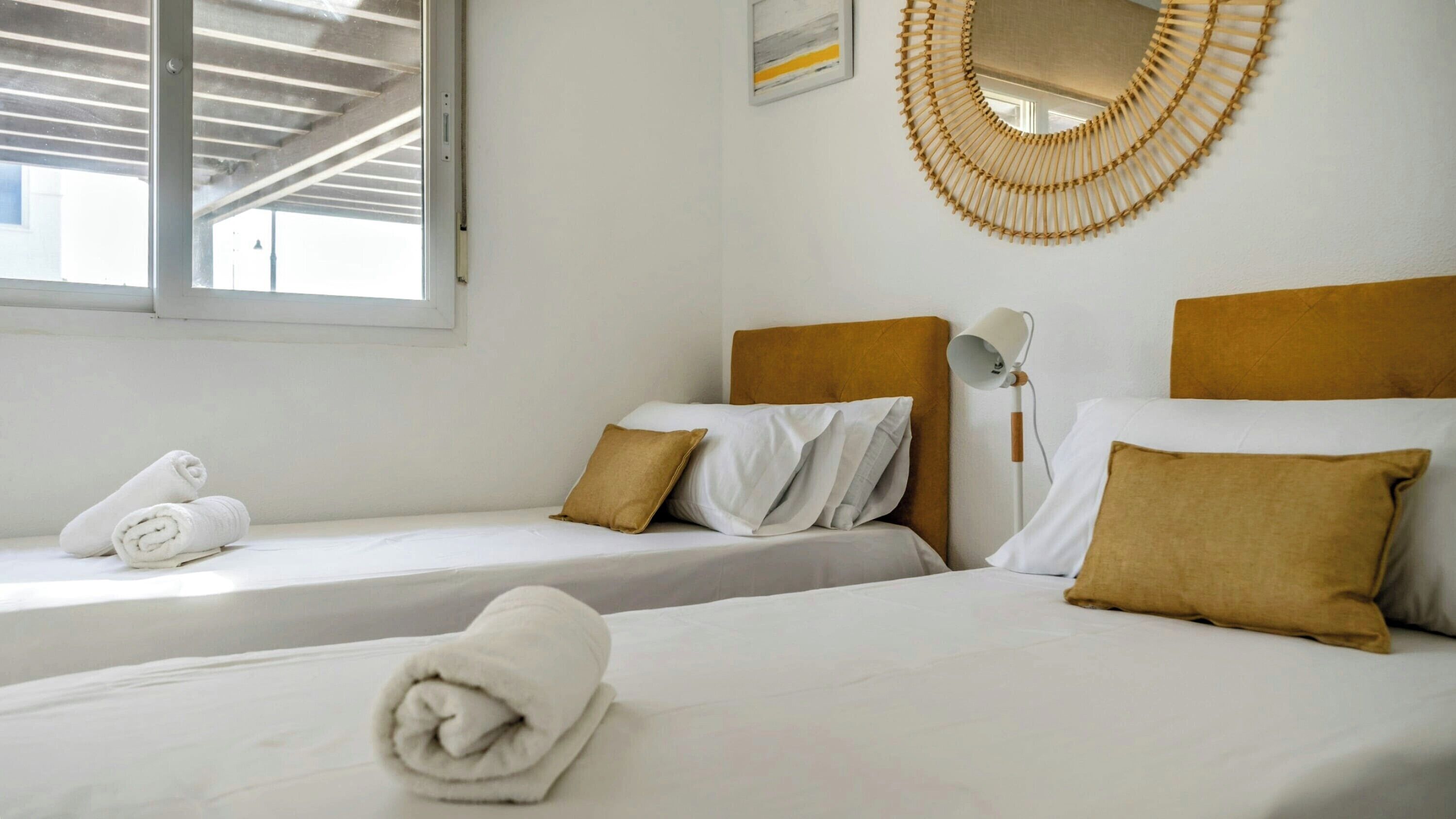 A hotel room with two single beds, white linens, brown pillows, a wall-mounted mirror, and a window with a view of the sky.