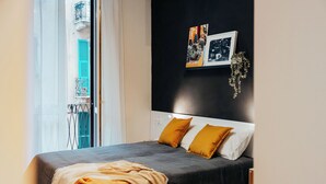 A modern bedroom with a large bed, two pillows, a bedside table, and a wall-mounted shelf with pictures and a plant.