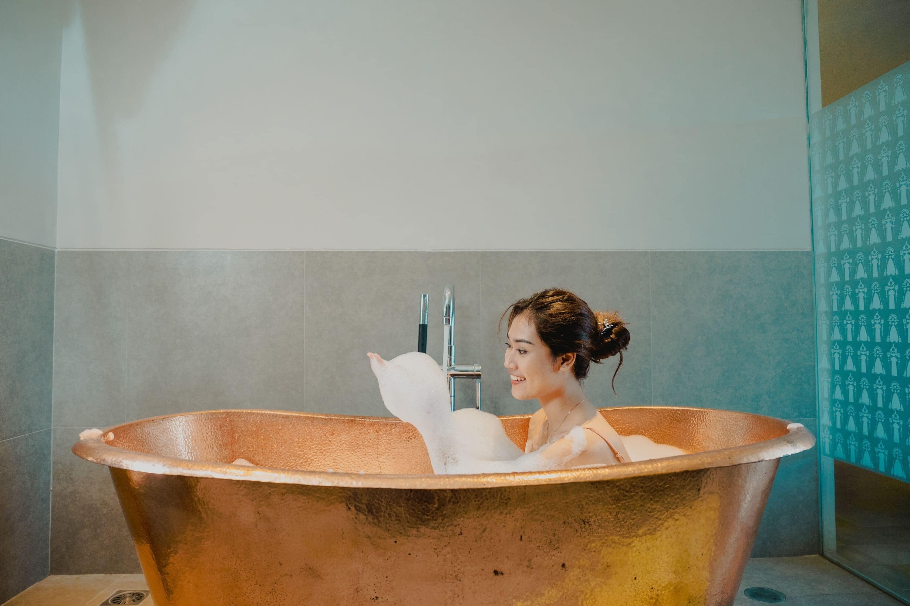 A person in a copper bathtub with a modern bathroom design, including grey tiles and a patterned wall.