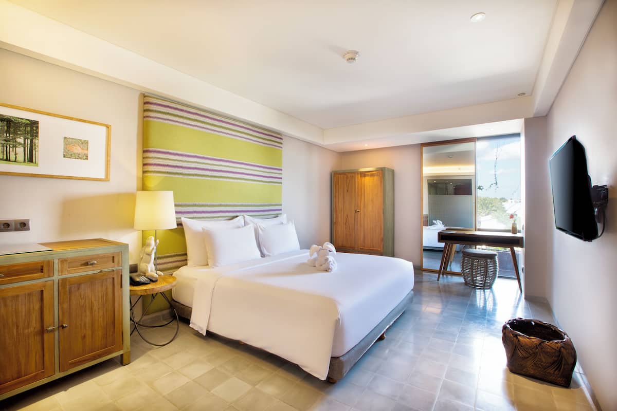 A hotel room with a large bed, a wooden wardrobe, a desk with a chair, a television mounted on the wall, and a window with a view.