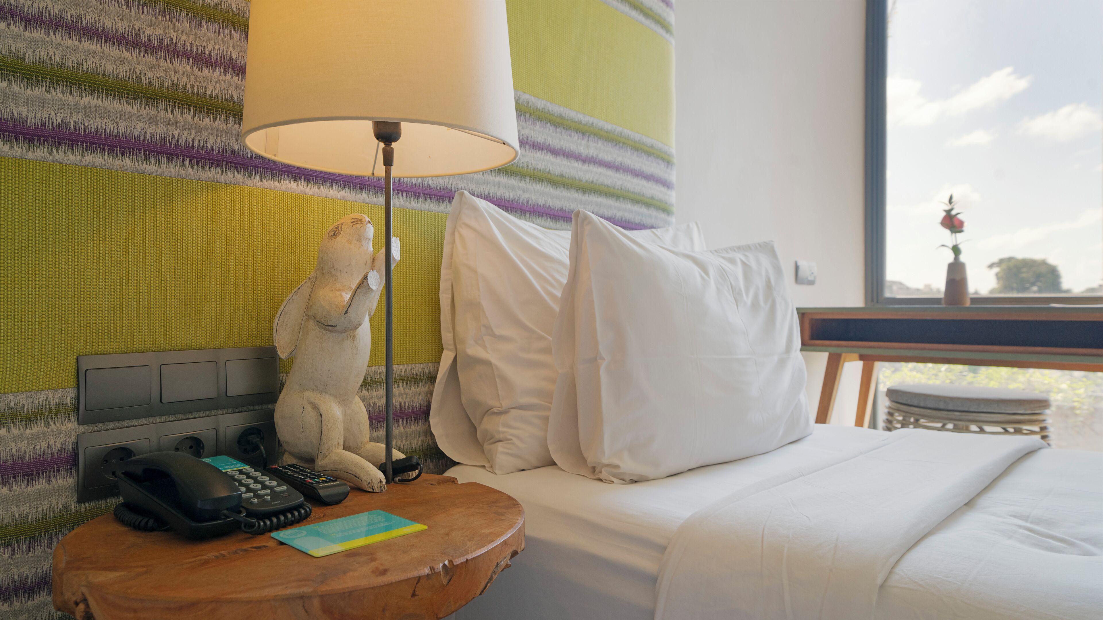 Une chambre d’hôtel avec un lit, une table de chevet avec un téléphone et des télécommandes, une lampe et une fenêtre donnant sur un arbre et un vase avec une fleur.