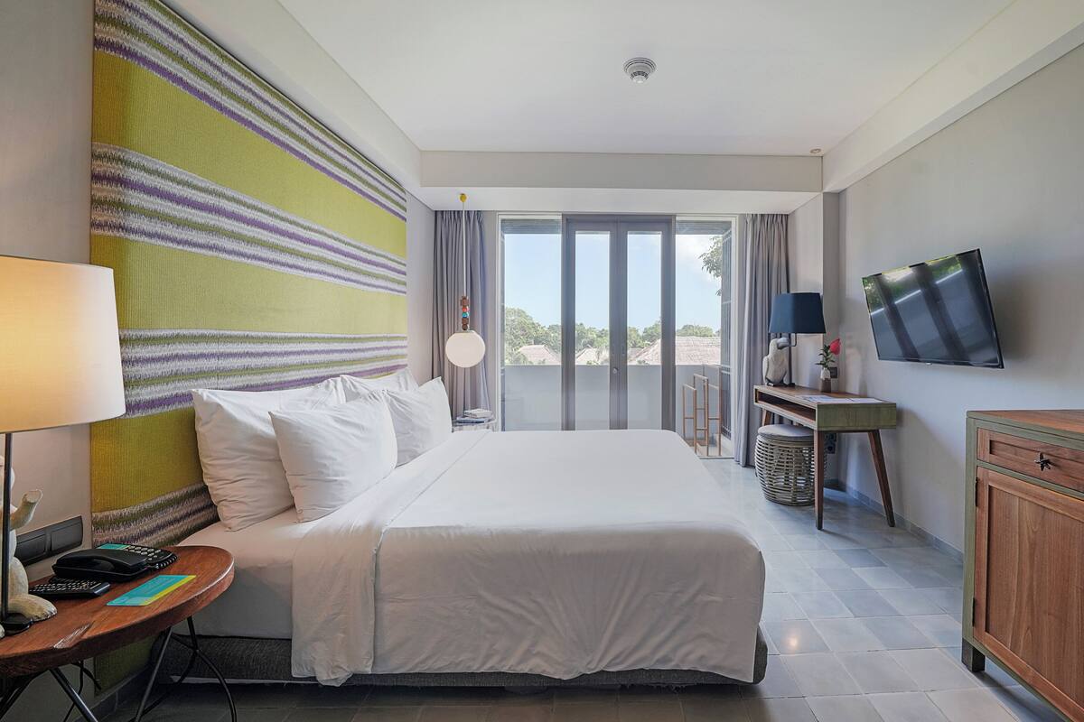 A hotel room with a bed, a desk with a lamp, a TV, and a view of the outdoors.