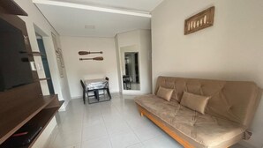 Un salon avec un canapé, un téléviseur posé sur un meuble, une table à manger avec des chaises, et un coin cuisine à l’arrière-plan.