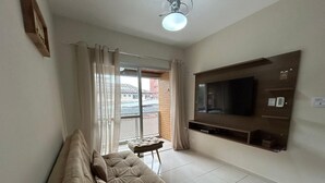 Un salon avec un canapé beige, un petit pouf, un téléviseur à écran plat fixé au mur et une porte-fenêtre coulissante donnant sur un balcon.