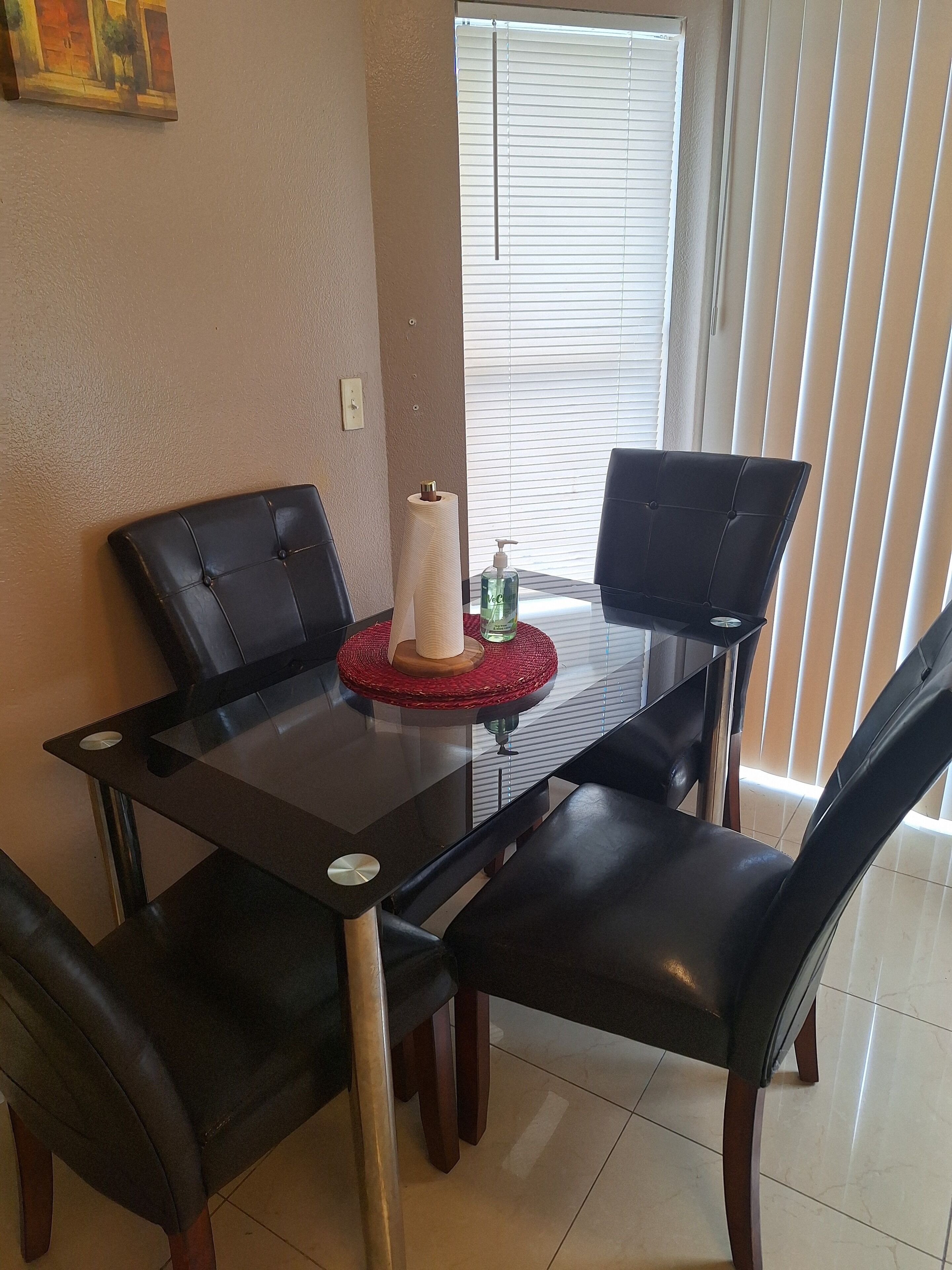Un coin repas avec une table en verre, des chaises en cuir noir, un set de table rouge, un flacon de gel hydroalcoolique et un tableau accroché au mur.