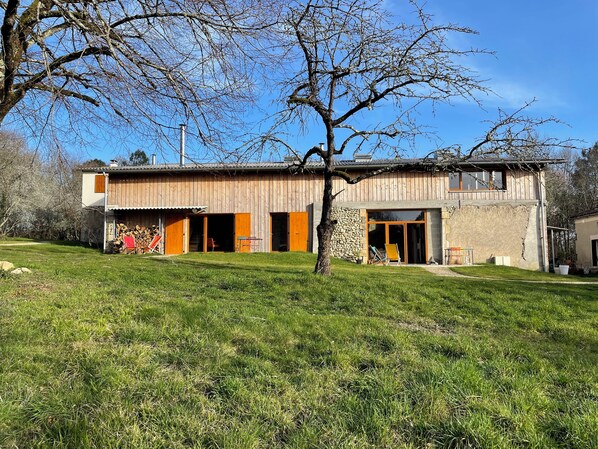 A rustic wooden house with a stone wall, a large open door, and a grassy yard.