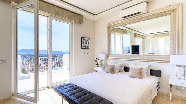 Ein Schlafzimmer mit einem großen Bett, einer Bank, einem Nachttisch, einem Spiegel und Blick auf die Stadt vom Balkon.