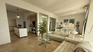 Ein modernes Wohnzimmer mit einem beigen Sofa, einem Glastisch und einem Essbereich mit weißem Tisch und Stühlen.